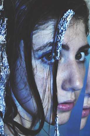 A close-up of a person's face with long dark hair partially covered by shiny metallic foil. The image is fragmented, creating a distorted and abstract look. The person has visible makeup and the focus is on their eyes and lips.