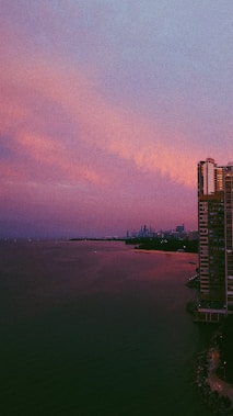 A high-rise building stands on the right, overlooking a body of water with a distant city skyline. The sky has hues of purple and pink, suggesting it is near sunrise or sunset.