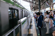 A group of people stand on a train platform, waiting as a train pulls into the station. Most individuals appear to be engaged with their phones, and there are platform markings for standing. The platform is covered by a roof and there are structural elements visible such as columns and signs.