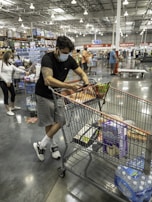 A person wearing a face mask is leaning on a shopping cart in a large warehouse-style store. The cart is filled with various items including water bottles and packaged goods. Other shoppers, also wearing masks, are seen in the background browsing the aisles. The interior is spacious with high ceilings and bright lighting.