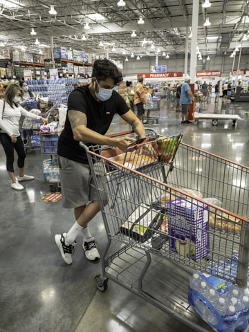 A person wearing a face mask is leaning on a shopping cart in a large warehouse-style store. The cart is filled with various items including water bottles and packaged goods. Other shoppers, also wearing masks, are seen in the background browsing the aisles. The interior is spacious with high ceilings and bright lighting.