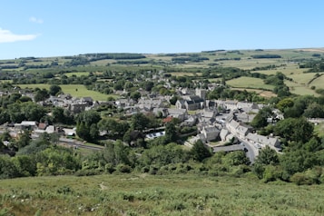 A picturesque village nestled amidst rolling green hills and farmland, featuring a cluster of stone buildings and a prominent church. Lush greenery and trees surround the area, creating a serene rural atmosphere under a clear blue sky.