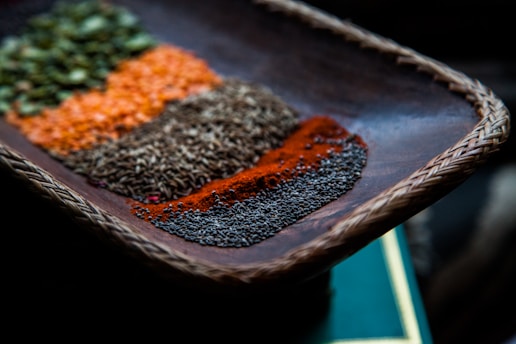 A close-up view of a woven wooden tray containing an assortment of colorful spices arranged in neat rows. The spices display a variety of textures and vibrant colors, including green, orange, brown, red, and black. The focus is on the details of each spice, highlighting their granular and powdered forms.