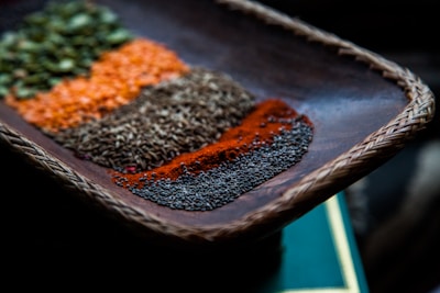 A close-up view of a woven wooden tray containing an assortment of colorful spices arranged in neat rows. The spices display a variety of textures and vibrant colors, including green, orange, brown, red, and black. The focus is on the details of each spice, highlighting their granular and powdered forms.