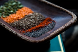 A close-up view of a woven wooden tray containing an assortment of colorful spices arranged in neat rows. The spices display a variety of textures and vibrant colors, including green, orange, brown, red, and black. The focus is on the details of each spice, highlighting their granular and powdered forms.