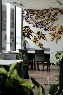 A modern dining area featuring a large wall mural with abstract foliage patterns in blue and yellow. The space is filled with natural light through large windows, and there are several green plants placed around the room. The dining table is surrounded by sleek chairs, creating a stylish and contemporary ambiance.