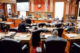 A legislative chamber with people sitting at wooden desks and chairs arranged in rows, surrounded by ornate architectural details. The room features red walls with decorative columns, a clock on the wall above the speaker's area, and several flags. Individuals are engaged in discussions and using laptops or reviewing documents.