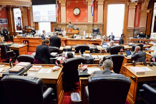 A legislative chamber with people sitting at wooden desks and chairs arranged in rows, surrounded by ornate architectural details. The room features red walls with decorative columns, a clock on the wall above the speaker's area, and several flags. Individuals are engaged in discussions and using laptops or reviewing documents.