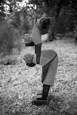 A black and white photograph features a sculpture composed of geometric shapes and rough stone, resembling an abstract human form bending forward. The background consists of a blurred natural setting with trees and foliage.