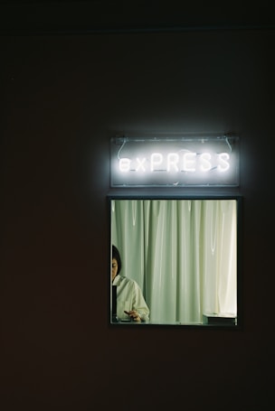 A dimly lit setting with a neon sign reading 'e x P R E S S' above a window or countertop. Through the window, a person is partly visible, standing behind a curtain while looking down, possibly working on something.