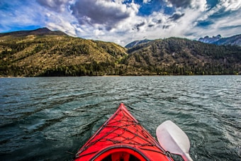 A vibrant red kayak is gliding across a large, serene lake, surrounded by lush, green mountains. The sky above is partly cloudy with varying shades of gray and blue.
