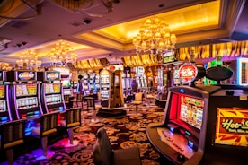 A vibrant casino interior filled with rows of colorful slot machines, each illuminated with bright lights and displays. The ceiling is adorned with elegant chandeliers and golden drapes, creating a luxurious atmosphere. The patterned carpet adds to the elaborate design of the space.