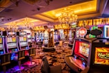 A vibrant casino interior filled with rows of colorful slot machines, each illuminated with bright lights and displays. The ceiling is adorned with elegant chandeliers and golden drapes, creating a luxurious atmosphere. The patterned carpet adds to the elaborate design of the space.