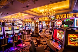 A vibrant casino interior filled with rows of colorful slot machines, each illuminated with bright lights and displays. The ceiling is adorned with elegant chandeliers and golden drapes, creating a luxurious atmosphere. The patterned carpet adds to the elaborate design of the space.