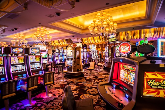 A vibrant casino interior filled with rows of colorful slot machines, each illuminated with bright lights and displays. The ceiling is adorned with elegant chandeliers and golden drapes, creating a luxurious atmosphere. The patterned carpet adds to the elaborate design of the space.