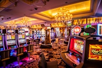 A vibrant casino interior filled with rows of colorful slot machines, each illuminated with bright lights and displays. The ceiling is adorned with elegant chandeliers and golden drapes, creating a luxurious atmosphere. The patterned carpet adds to the elaborate design of the space.