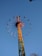 A colorful amusement park ride extends into the sky, resembling a giant swing with seats attached to long arms radiating from a central column. People are seated, enjoying the thrill as the ride spins in a circular motion against a clear blue sky.