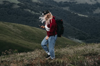 A person with long hair wearing a red sweater, jeans, and a black backpack is standing on a grassy hillside, looking at a smartphone. The background features a vast landscape of rolling green hills and dense forest.