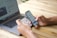 A person is holding a smartphone and a credit card, engaged in an online payment transaction. A laptop with a webpage open is positioned on a wooden table.