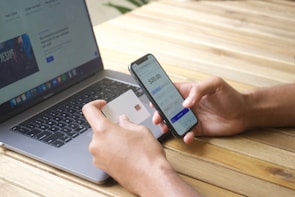 A person is holding a smartphone and a credit card, engaged in an online payment transaction. A laptop with a webpage open is positioned on a wooden table.