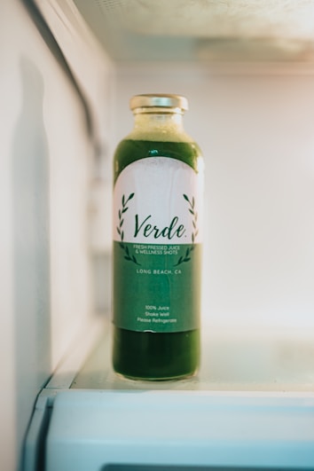 A bottle of green juice labeled 'Verde' sits inside a refrigerator. The label indicates it is fresh-pressed juice and wellness shots from Long Beach, CA. The clear bottle allows the green liquid to be visible, and the top is sealed with a metallic cap.