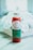 A glass bottle filled with red liquid, labeled 'Verde' with details about fresh pressed juice and wellness shots from Long Beach, CA, sits on a surface. The label is green and white, and the bottle is sealed with a metallic cap. In the background, there is a blurred view of white flowers.