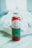 A glass bottle filled with red liquid, labeled 'Verde' with details about fresh pressed juice and wellness shots from Long Beach, CA, sits on a surface. The label is green and white, and the bottle is sealed with a metallic cap. In the background, there is a blurred view of white flowers.