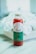 A glass bottle filled with red liquid, labeled 'Verde' with details about fresh pressed juice and wellness shots from Long Beach, CA, sits on a surface. The label is green and white, and the bottle is sealed with a metallic cap. In the background, there is a blurred view of white flowers.