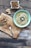 A bowl of green soup with a swirl of cream and topped with seeds placed on a rustic wooden table. Nearby, there is a wooden board with a spoonful of crushed chili and chopped mushrooms, as well as a piece of ginger. In the corner, a small dish contains a dry crumbly mixture with a wooden spoon.