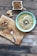 A bowl of green soup with a swirl of cream and topped with seeds placed on a rustic wooden table. Nearby, there is a wooden board with a spoonful of crushed chili and chopped mushrooms, as well as a piece of ginger. In the corner, a small dish contains a dry crumbly mixture with a wooden spoon.