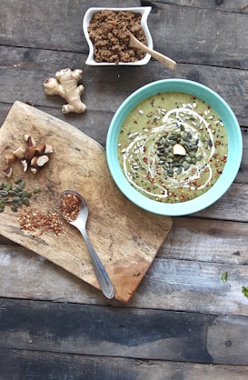 A bowl of green soup with a swirl of cream and topped with seeds placed on a rustic wooden table. Nearby, there is a wooden board with a spoonful of crushed chili and chopped mushrooms, as well as a piece of ginger. In the corner, a small dish contains a dry crumbly mixture with a wooden spoon.