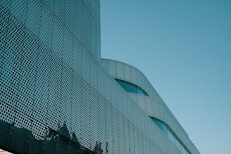 A modern architectural structure features a facade with a perforated metal design. The curved lines and metallic texture create a futuristic appearance against a clear blue sky.
