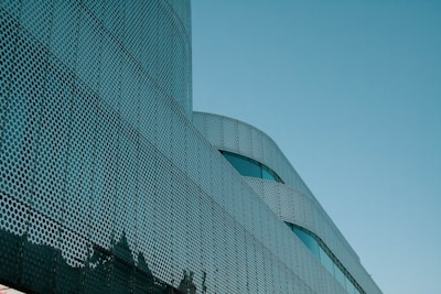 A modern architectural structure features a facade with a perforated metal design. The curved lines and metallic texture create a futuristic appearance against a clear blue sky.