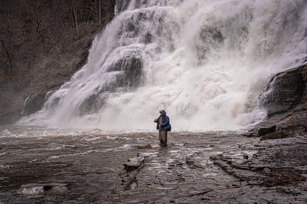 Hombre con chaqueta azul y pantalones cortos negros parado en la roca cerca de las cascadas durante el día