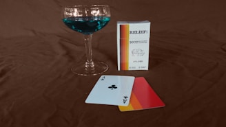 A cocktail glass filled with a vibrant blue liquid stands next to a pack of CBD cigarettes. Two playing cards, an ace of clubs and an ace with a gradient design, lie on a dark brown surface.