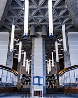 A modern interior featuring tall cylindrical lights hanging from a patterned ceiling. The architecture has a geometric, industrial style with concrete elements and a central pillar. Below, there are curved glass and wooden panels, creating a symmetrical design.