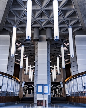 A modern interior featuring tall cylindrical lights hanging from a patterned ceiling. The architecture has a geometric, industrial style with concrete elements and a central pillar. Below, there are curved glass and wooden panels, creating a symmetrical design.