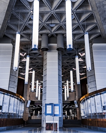 A modern interior featuring tall cylindrical lights hanging from a patterned ceiling. The architecture has a geometric, industrial style with concrete elements and a central pillar. Below, there are curved glass and wooden panels, creating a symmetrical design.