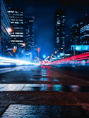 Dark cityscape with neon red and white lights reflecting on wet streets.