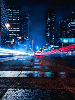 Dark cityscape with neon red and white lights reflecting on wet streets.