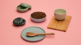 A minimalist arrangement of kitchenware items on a pastel pink background, including a green ceramic dish with a wooden spoon, a ribbed green cup on a bamboo board, a lidded green ceramic container, and a bowl filled with coffee beans.