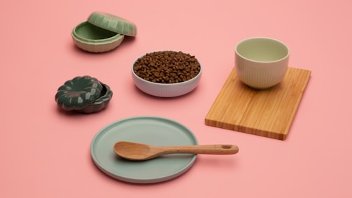 A minimalist arrangement of kitchenware items on a pastel pink background, including a green ceramic dish with a wooden spoon, a ribbed green cup on a bamboo board, a lidded green ceramic container, and a bowl filled with coffee beans.