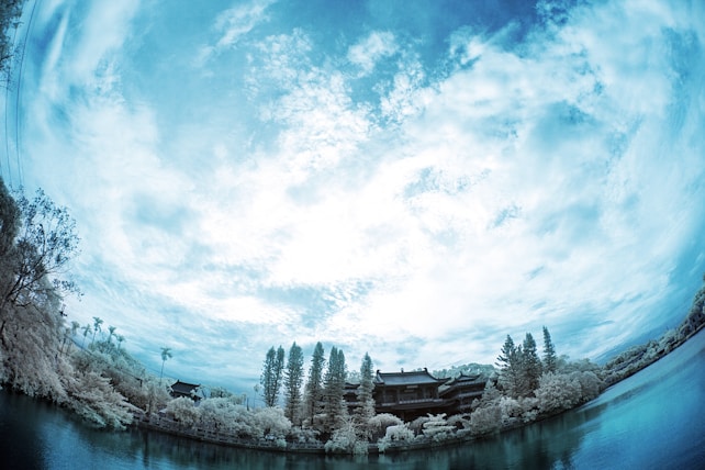 A serene landscape features a large expanse of sky filled with wispy clouds. Below, a still body of water reflects the scene, bordered by a variety of trees and traditional-style structures nestled among the foliage. The image is captured with an infrared filter, giving it a cool blue and white tone.