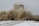 A large cloud of dust billows from a collapsing structure while two people wearing protective gear appear to be moving away from the scene. The ground is covered with snow, and debris is scattered around.