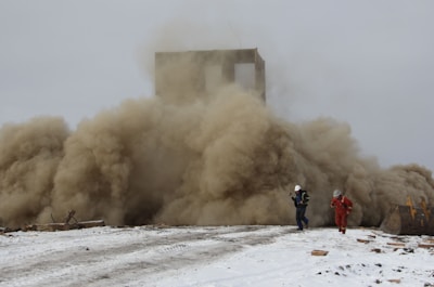 A large cloud of dust billows from a collapsing structure while two people wearing protective gear appear to be moving away from the scene. The ground is covered with snow, and debris is scattered around.