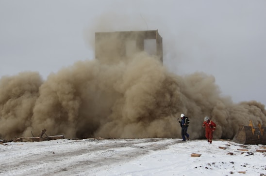 A large cloud of dust billows from a collapsing structure while two people wearing protective gear appear to be moving away from the scene. The ground is covered with snow, and debris is scattered around.
