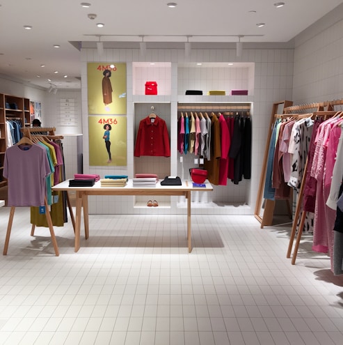 A neatly organized clothing store featuring a variety of colorful clothing items displayed on hangers and shelves. A central wooden table showcases folded garments with vibrant colors. The walls have posters and more hanging clothes. The store has a modern, minimalist aesthetic with a bright and clean interior.