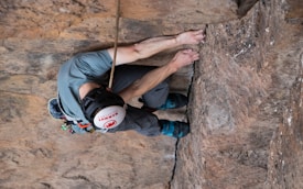 A climber grips the rough surface of a rock wall, using hands and feet to navigate an overhang. They are wearing a helmet, climbing shoes, and a harness with gear attached. The rock appears rugged and textured.
