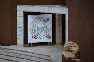 A modern architectural entrance with a large colorful poster featuring abstract shapes and text. The area includes stone steps leading up to the entrance and a sculpted stone bust on the right. The building facades are clad in brown tiles, creating a minimalistic yet artistic atmosphere.