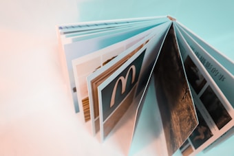 An open, colorful photo book with several pages displaying images, viewed from an angled side perspective. The pages appear to contain photographs and possibly text, with one page prominently featuring a logo that resembles an 'M'. The overall color scheme is light with a soft focus in parts of the image.